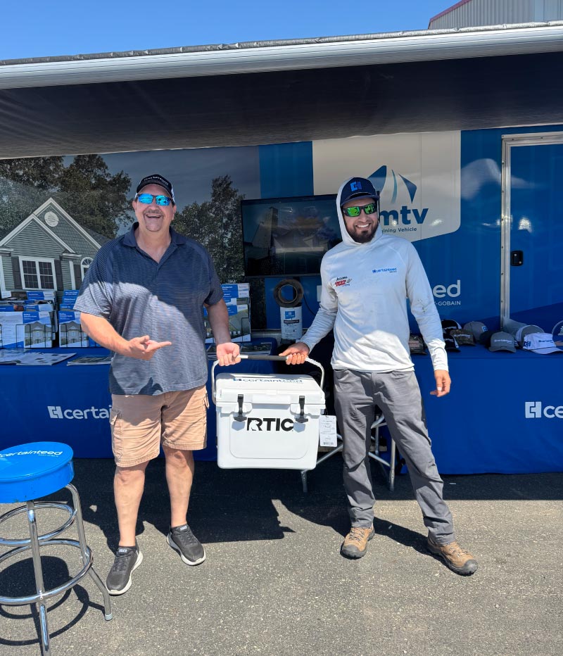 roofers two roofers from dakota storm damage standing outdoors in front of a promotional setup, holding an RTIC cooler between them.