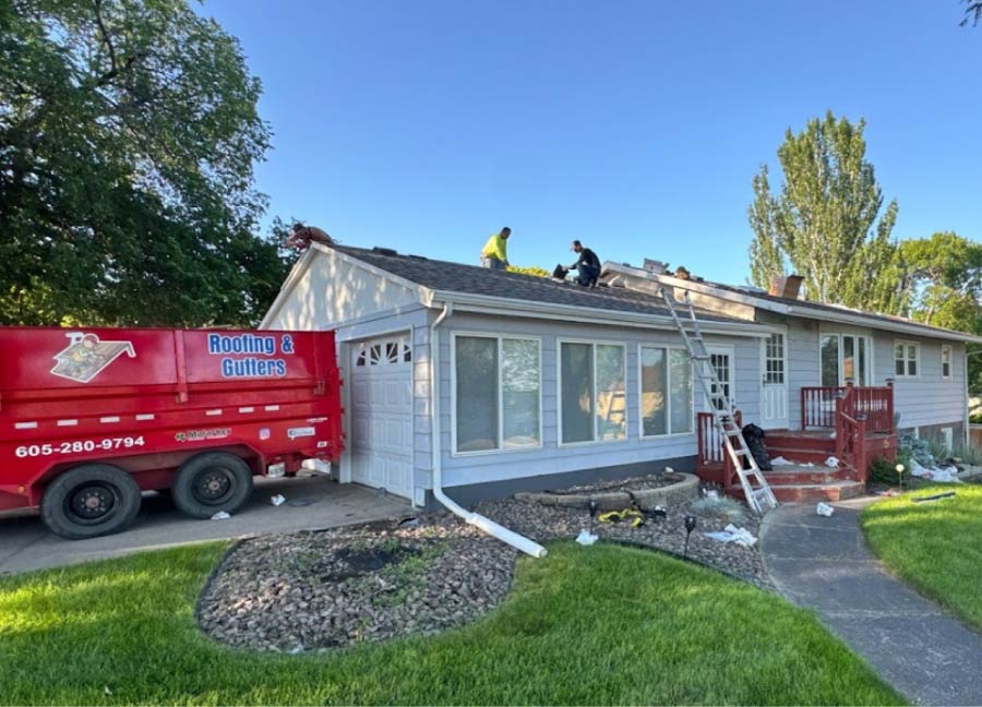 roofing-crew Dakota Storm Roofing crew replacing asphalt shingles on a residential home using an Equipter debris trailer to protect the property during storm restoration services.