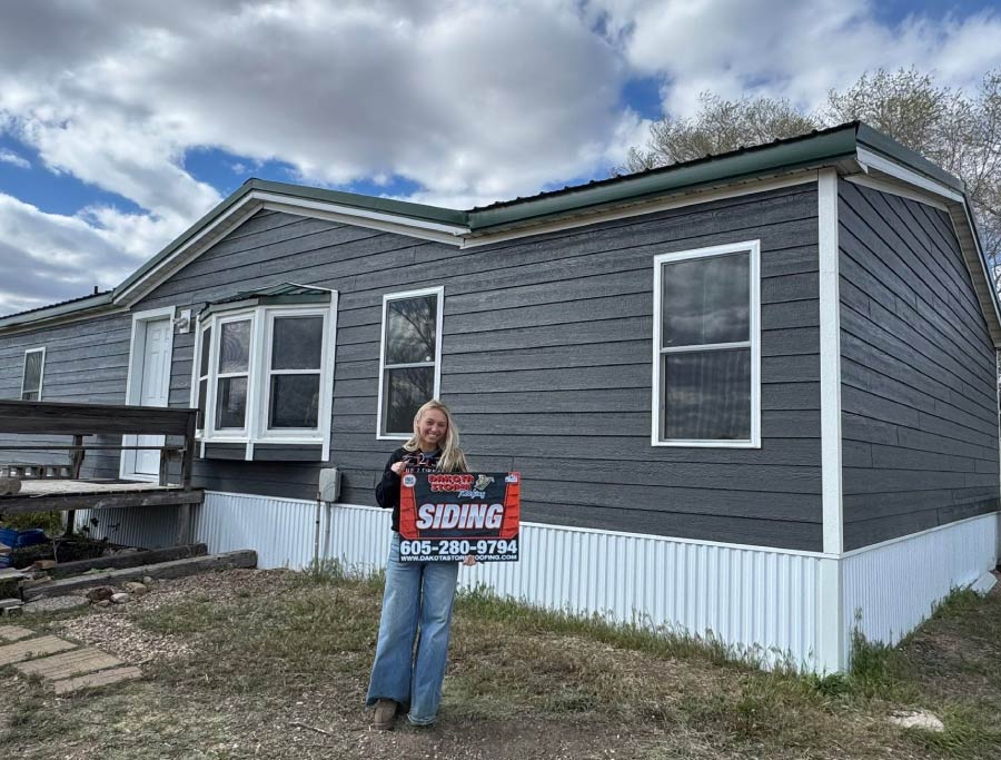 siding-project A happy customer standing in front of a home with newly installed dark grey siding, holding a Dakota Storm Roofing sign with our contact information in Pierre, South Dakota.